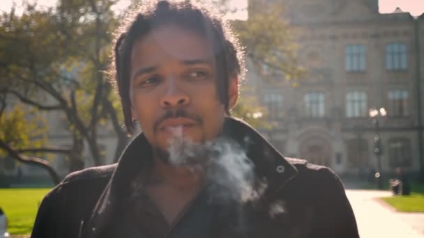 Nahaufnahme Porträt eines afrikanisch-amerikanischen Typen mit Dreadlocks, der auf herbstlichem Campus-Hintergrund raucht. — Stockvideo