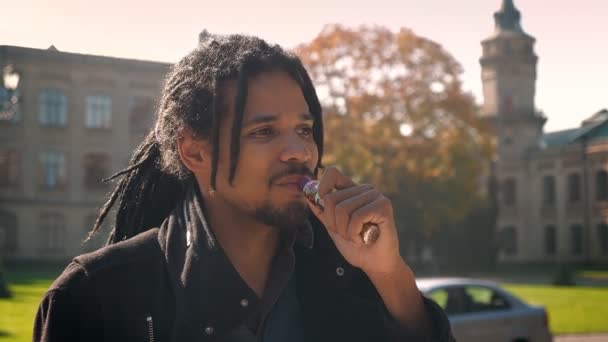 Closeup portret van Afro-Amerikaanse man met dreadlocks roken en kijken in de camera op herfst campus achtergrond. — Stockvideo