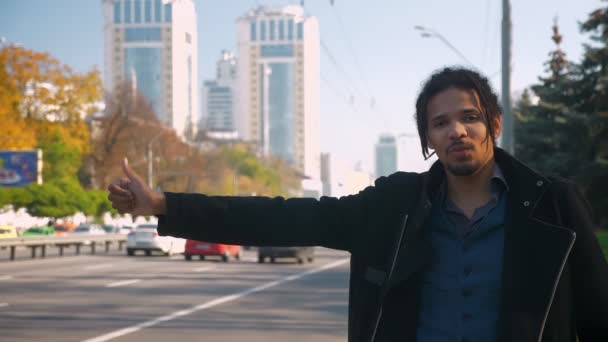 Portrait of tired African-American guy with dreadlocks nervously hitchhiking on road background. — Stock Video