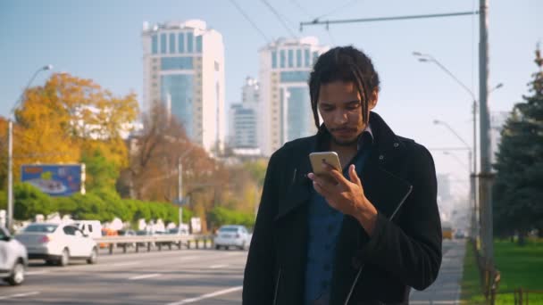 Portrait de gars afro-américain fatigué avec dreadlocks nerveusement debout près de la route avec téléphone dans les mains . — Video