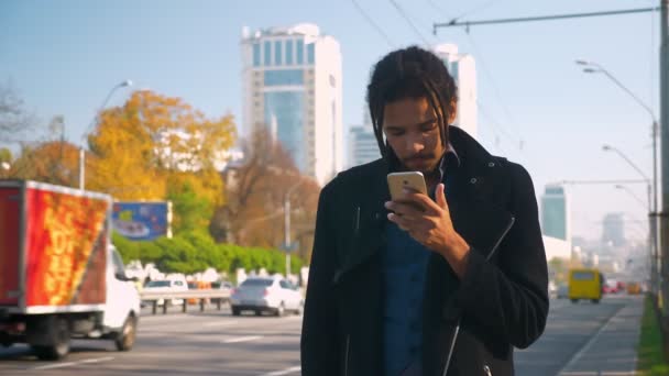 Portrait of tired African-American guy with dreadlocks nervously standing near road and watching into smartphone. — Stock Video