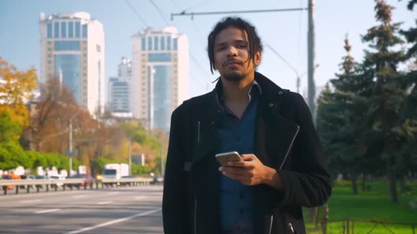 Retrato de un joven afroamericano con rastas mirando hacia el smartphone y mirando nerviosamente hacia la carretera . — Vídeos de Stock