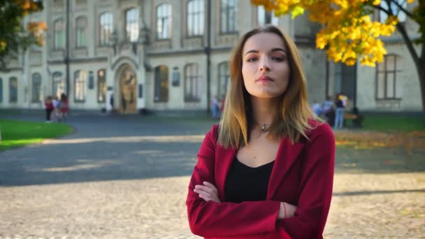 Jonge vrouwelijke student met gekruiste handen vol vertrouwen op zoek op een camera op de achtergrond van de Universiteit — Stockvideo
