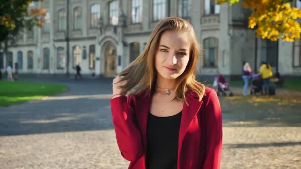 Young female student smiling and turning her head looking on a camera, playing with her hair, on the background of university — Stock Video