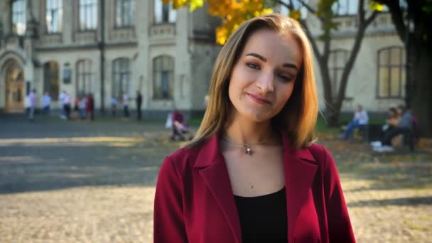 Jovem mulher atraente, estudante inclinando a cabeça à esquerda, sorrindo, levantando a sobrancelha no fundo da universidade, ao ar livre em um dia ensolarado — Vídeo de Stock