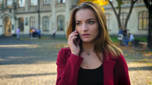 Ung kvinnlig student berättar på sin telefon utanför universitetet, blir upprörd och ser på klockan. Väntar utanför universitetet — Stockvideo