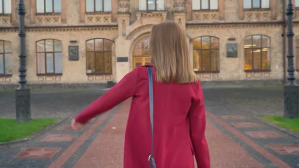 Filmagem de uma jovem estudante caminhando até a entrada da universidade acenando com a mão, cumprimentando, pelas costas — Vídeo de Stock