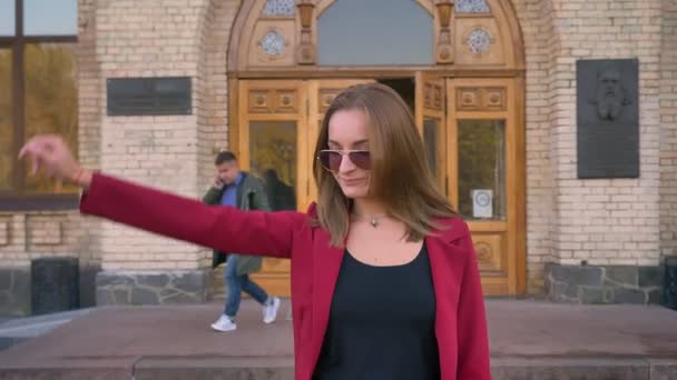 Giovane studentessa attraente che esce dall'università, salutando qualcuno e sorridendo cammina per la sua strada — Video Stock