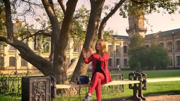 Stunning young female student sitting in the university square and taking selfies, perceiving nature in a sunny warm day — Stock Video