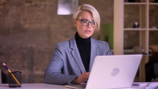 Портрет короткошерстої бізнес-леді в окулярах, що працюють з ноутбуком в офісі і дивиться в камеру з посмішкою . — стокове відео