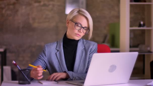 Portret van blonde korthaar zakenvrouw in glazen werken met de laptop en het maken van aantekeningen in het notitieblok in office. — Stockvideo