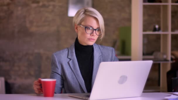 戴眼镜的金发碧眼的女商人的肖像 她用心工作 在办公室里用笔记本电脑和喝咖啡 — 图库视频影像