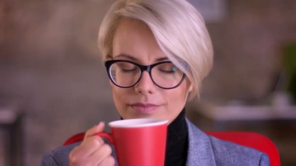 Porträt der blonden, kurzhaarigen Geschäftsfrau mit Brille, die einladend in die Kamera schaut und im Büro Kaffee trinkt. — Stockvideo