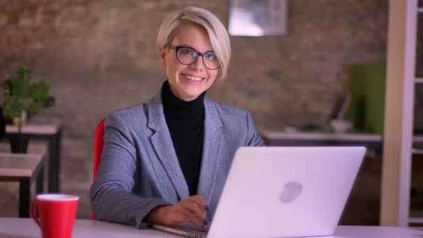 Retrato de mujer de negocios rubia de pelo corto en gafas trabajando con laptop y logrando éxitos felizmente en oficina . — Vídeo de stock