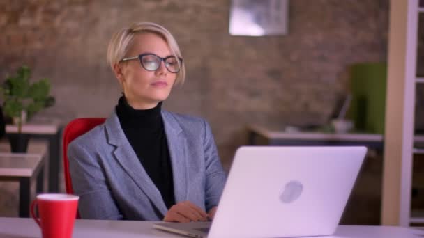 Portret van blonde korthaar zakenvrouw in glazen aandachtig kijken naar de laptop en drinken koffie in office. — Stockvideo