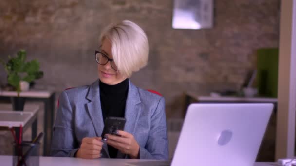 Портрет короткошерстої бізнес-леді в окулярах, яка дивиться в смартфон і сміється в офісі . — стокове відео