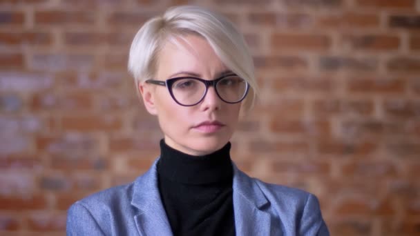 Portrait de femme blonde aux cheveux courts d'âge moyen dans des lunettes regardant sérieusement à la caméra sur fond de mur de poulet . — Video