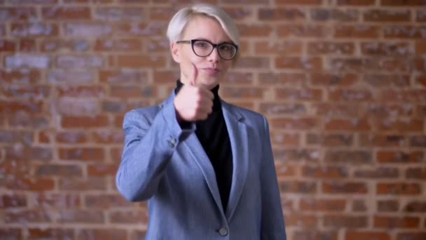 Retrato de mujer rubia caucásica de pelo corto en gafas haciendo gestos con el dedo hacia arriba para mostrar como sobre fondo de pared de ladrillo . — Vídeos de Stock