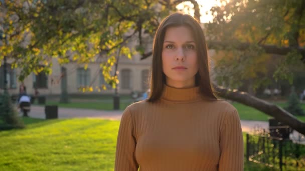 Retrato de una joven morena caucásica mirando en serio a la cámara sobre fondo verde del parque . — Vídeos de Stock