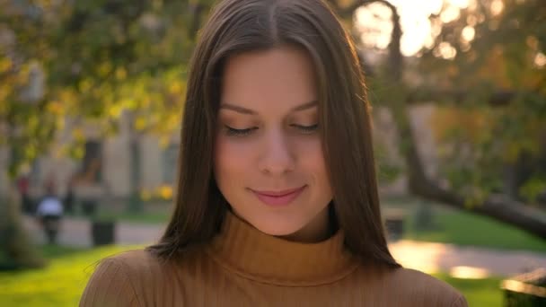 Крупный план портрета юной брюнетки, скромно избегающей зрительного контакта с камерой на фоне зеленого парка . — стоковое видео
