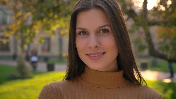 Primer plano retrato de la joven morena mirando a la cámara, coqueteando y sonriendo sobre el fondo verde del parque . — Vídeo de stock