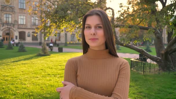 Ritratto di giovane ragazza bruna caucasica che guarda allegramente nella fotocamera con le braccia incrociate sullo sfondo del parco verde . — Video Stock