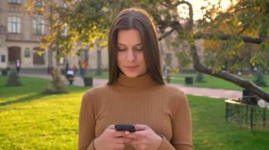 Güzel beyaz kız sörf ve sohbet smartphone kullanarak ve duygular üzerinde yeşil park arka plan değiştirme.