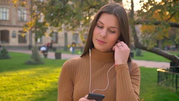 Portret piękna brunetka, stawiając na słuchawki i tańczyć do muzyki na tle zielonego parku. — Wideo stockowe