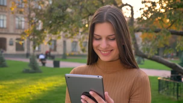 Mooie brunette surfen op de tablet en glimlachend op groen park achtergrond. — Stockvideo
