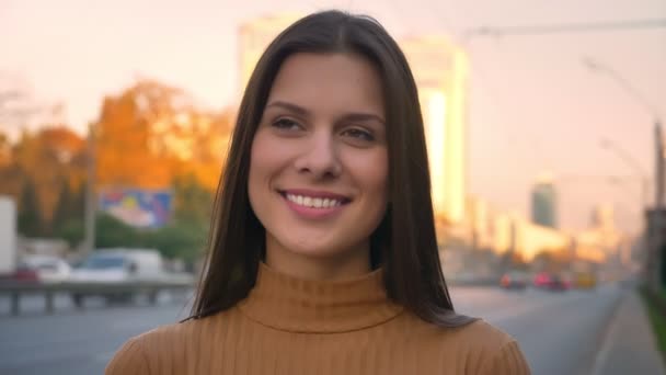 Close-up retrato de menina morena atraente se voltando para câmera wih sorriso sincero no fundo da estrada — Vídeo de Stock