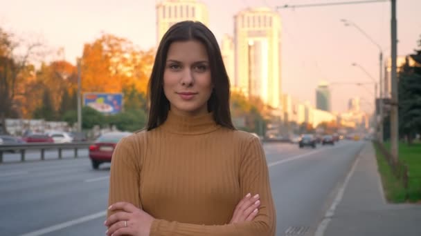 Portrait of serious caucasian brunette watching modestly into camera with her arms crossed on road background. — Stock Video