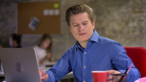 Portrait of middle-aged blond caucasian businessman typing on laptop in office, drinking coffee and smiling to camera. — Stock Video