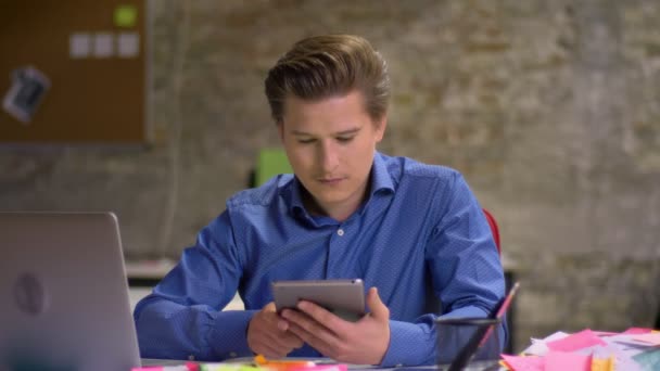 Portrait of middle-aged blond businessman works in office using tablet and shows its display vertically to camera. — Stock Video