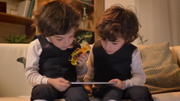 Portrait de petits jumeaux arabes bouclés assis sur le canapé et regardant avec intérêt dans la tablette dans le salon . — Video