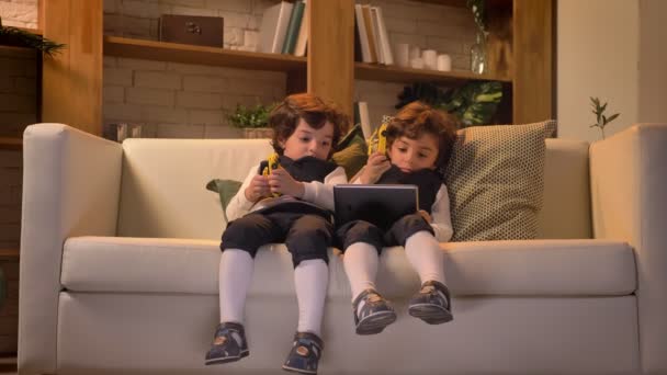 Портрет довольно арабских кудрявых близнецов, сидящих на диване и интересно смотрящих на планшет в гостиной . — стоковое видео