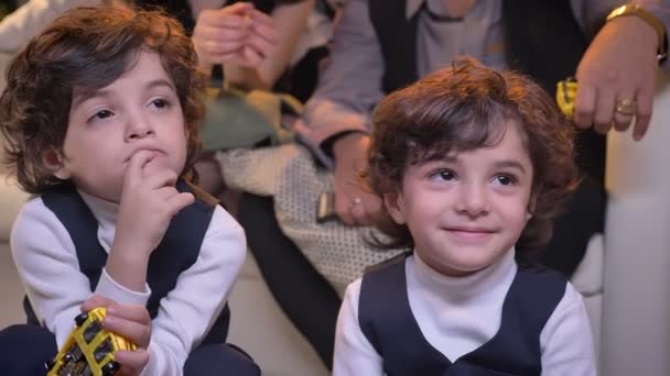 Två ganska arabiska lockigt-headed tvillingar sitter på golvet i vardagsrummet leende titta på Tv med sin familj på bakgrund. — Stockvideo