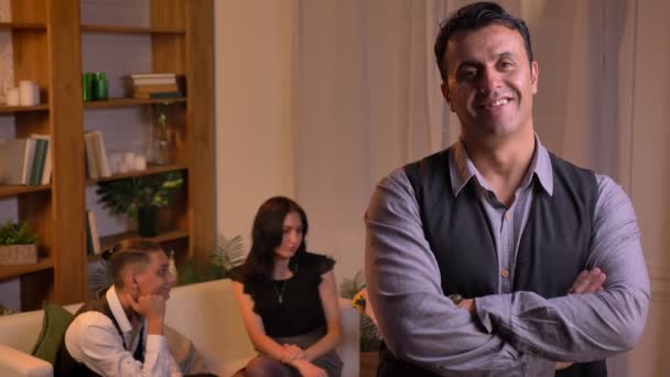 Retrato del hombre árabe mirando sonriente a cámara y brazos cruzados de pie en la sala de estar con la familia en el fondo . — Vídeos de Stock