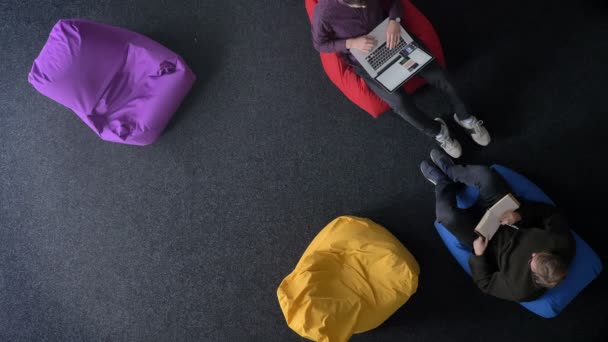 Top-shot van twee jonge mannen die werken met de laptop en notebook op zachte veelkleurig tassen snel veranderende zetels. — Stockvideo