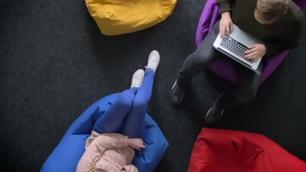 Circling around topshot of man working with laptop and woman talking on cellphone sitting on multicoloured bags. — Stock Video