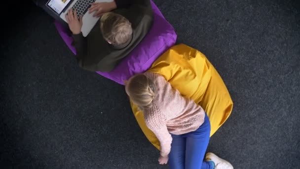 Circulando em torno de topshot de homem que trabalha com laptop e mulher curiosamente se voltando para ele sentado em sacos multicoloridos . — Vídeo de Stock