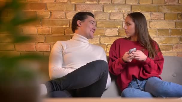 Улыбающаяся молодая кавказская пара сидит на диване, смотрит в смартфон и разговаривает друг с другом в домашней атмосфере . — стоковое видео