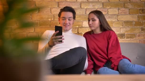 Casal caucasiano sentado no sofá assistindo em smartphone com concentração e conversando uns com os outros em ambiente doméstico . — Vídeo de Stock