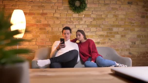 Junges kaukasisches Paar sitzt auf Sofa und schaut in Smartphone und kommentiert den Inhalt in häuslicher Atmosphäre. — Stockvideo