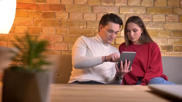 Young caucasian couple sitting on sofa attentively watching into tablet in home atmosphere. — Stock Video