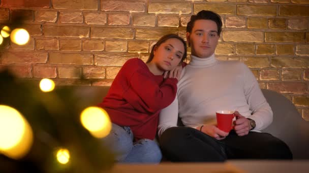 Портрет расслабляющей обнимающей кавказской пары, сидящей на диване и смотрящей кино с чашкой напитка в уютной домашней обстановке . — стоковое видео