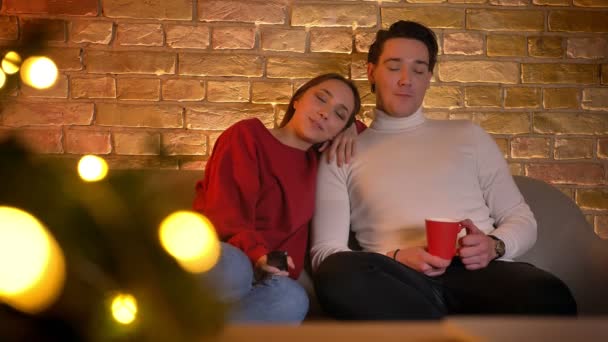 Sonriendo abrazando pareja caucásica sentado en el sofá y la elección de películas en la televisión con la taza de bebida en ambiente acogedor hogar . — Vídeos de Stock
