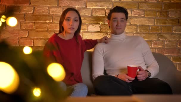 Abraçando casal caucasiano sentado no sofá e assistindo atentamente filme com xícara de bebida na atmosfera de Natal . — Vídeo de Stock
