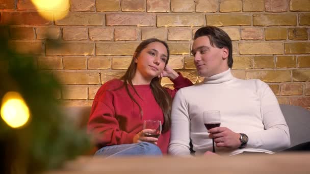 Retrato de pareja caucásica feliz sentada en el sofá y bebiendo vino hablando entre sí en un ambiente acogedor de Navidad . — Vídeo de stock