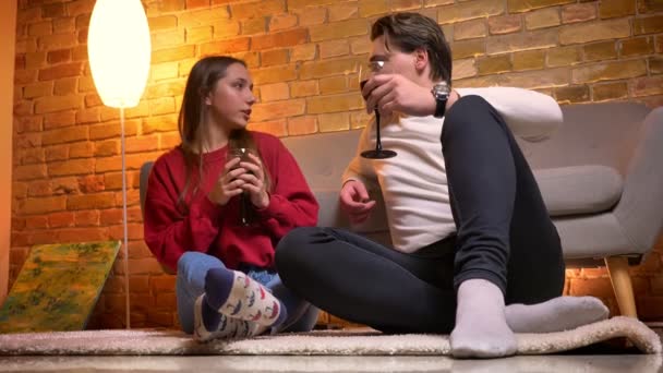 Close-up portrait of young caucasian friends sitting on carpet and drinking wine talking with each other in cosy home atmosphere. — Stock Video