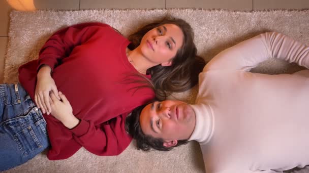 Top shot de jovem casal caucasiano deitado cabeça a cabeça no chão e conversando uns com os outros em atmosfera acolhedora . — Vídeo de Stock